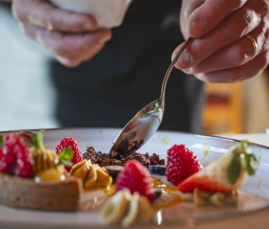 Una persona prepara un dessert su un piatto bianco, con una cucchiaio sopra una crostata e cioccolato sbriciolato a lato.