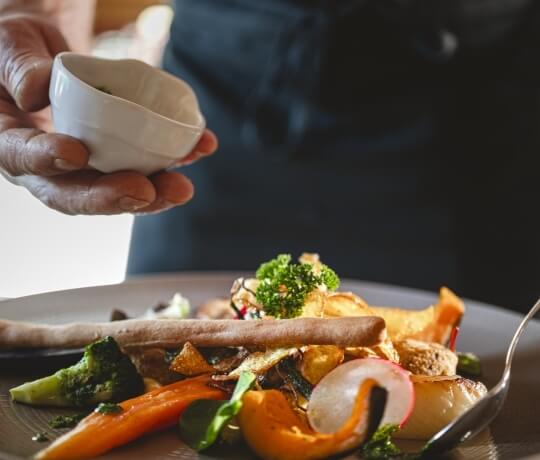 Una mano tiene un ramekin bianco con condimento sopra un piatto di verdure colorate in un ambiente di ristorante.