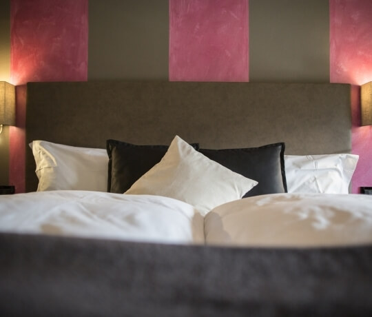 Camera d'hotel accogliente con letto, coperta bianca, cuscini bianchi e neri, lampade calde e tonalità rilassanti.