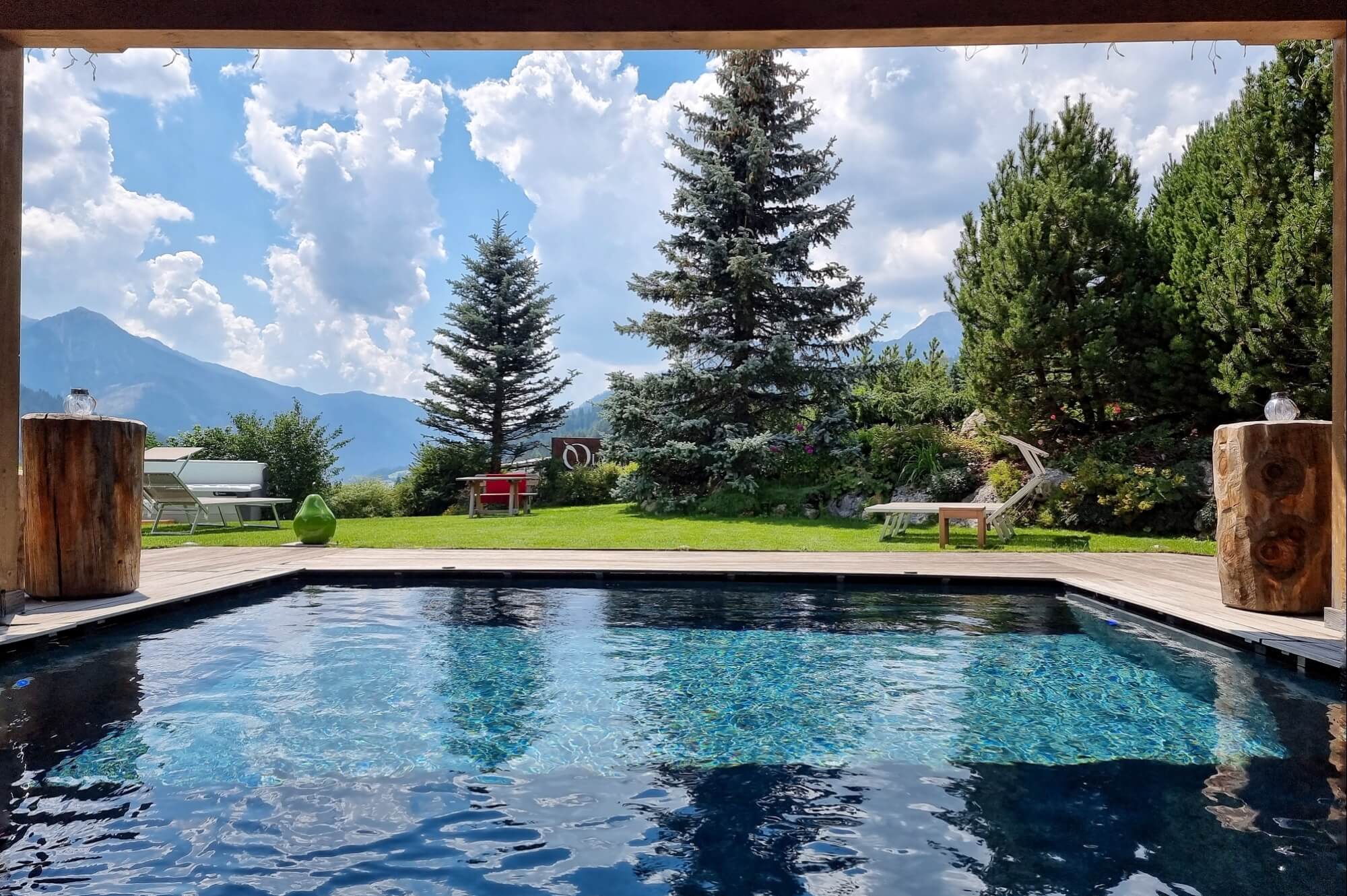 Immagine di una piscina circondata da un deck in legno, alberi, mobili da giardino e un paesaggio montano sullo sfondo.