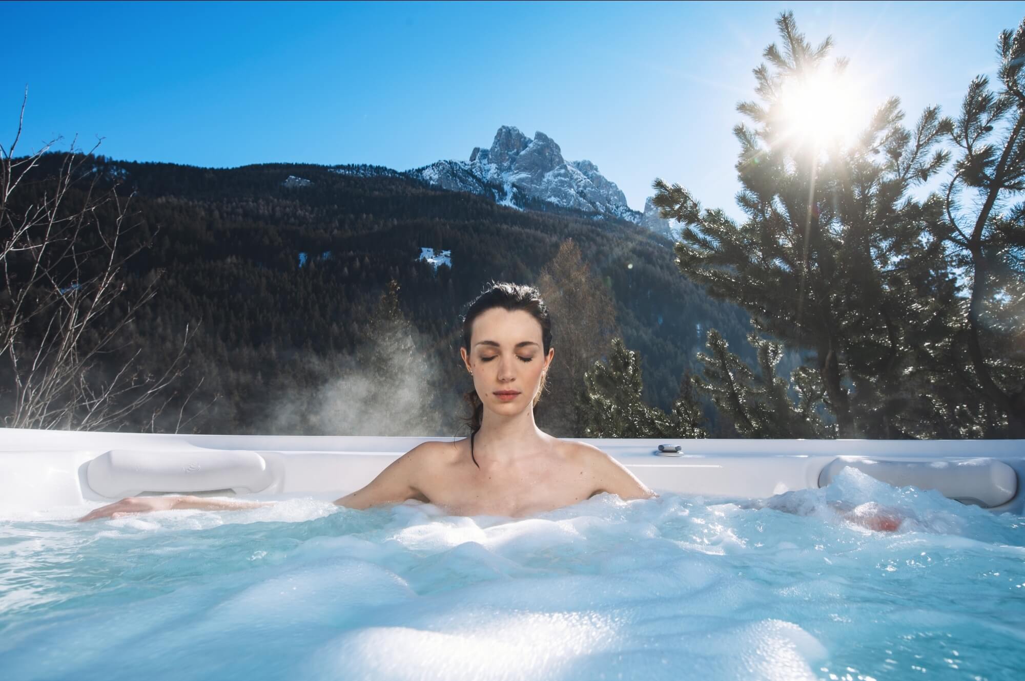 Donna serena in una vasca idromassaggio, circondata da montagne innevate e alberi, mentre medita sotto un cielo blu.
