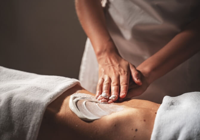 Una persona sdraiata sulla pancia mentre un'altra applica crema sulla schiena in un ambiente di spa.