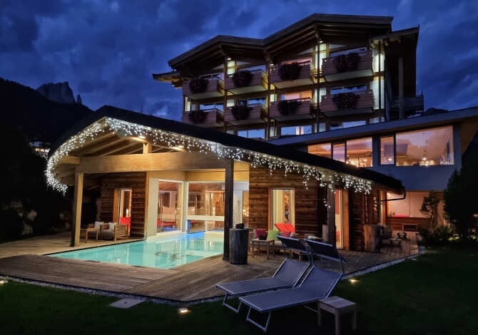 Casa moderna a più livelli con piscina, sedie a sdraio e luci bianche, circondata da un prato verde e cielo nuvoloso.