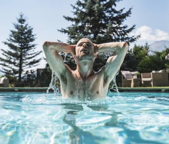 "Uomo in piscina con le braccia alzate, circondato da alberi e cielo azzurro."