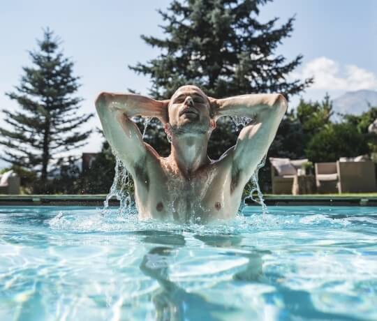 Uomo sdraiato in una piscina all'aperto, godendosi il sole con le braccia sollevate e alberi sullo sfondo.