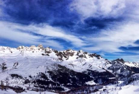 Paesaggio montano innevato con cielo blu, nuvole bianche e alberi verdi, evocando un'atmosfera serena invernale.