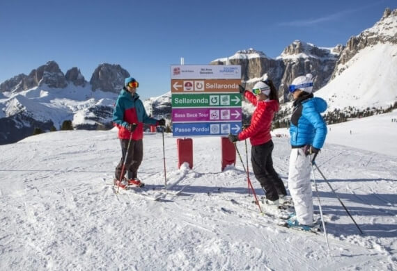 Tre sciatori colorati con attrezzatura in un paesaggio innevato, davanti a un cartello per Sellaronda e Passo Pordo.
