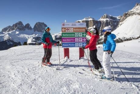 Tre persone in abbigliamento invernale con sci su una montagna innevata, davanti a un cartello con direzioni e distanze.