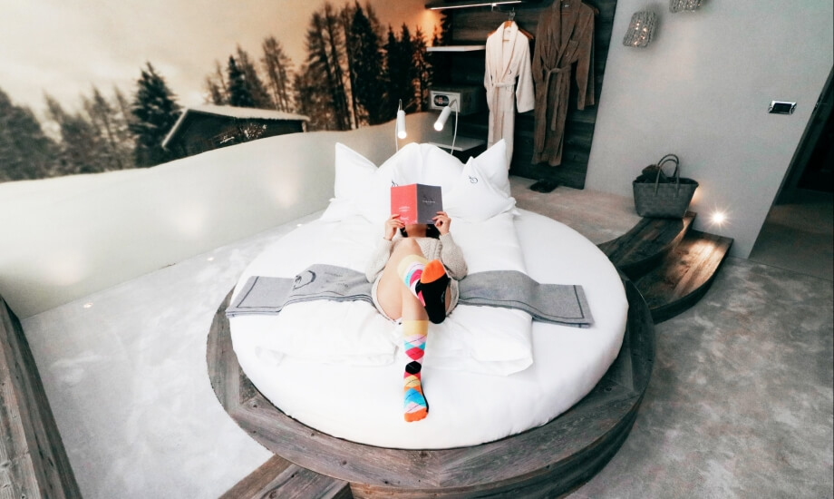 Una persona sdraiata su un letto circolare, immersa nella lettura di un libro, in una stanza serena con vista sul tramonto.