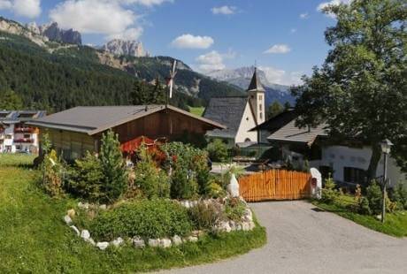 Un villaggio sereno tra le montagne, con chiesa, case, giardino fiorito e un panorama montano tranquillo.