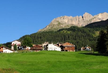 Villaggio sereno in montagna con edifici rustici, foresta verde e un picco roccioso sotto un cielo blu.