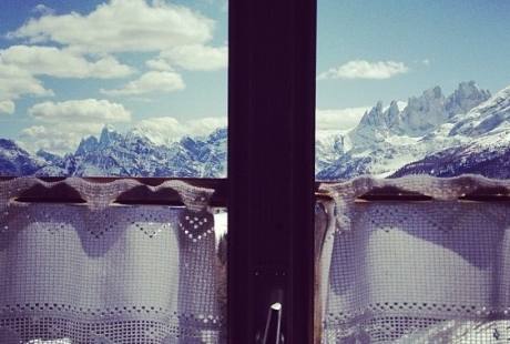 Vista di una catena montuosa attraverso una finestra con telaio marrone scuro, parzialmente coperta da una tenda di pizzo bianco.
