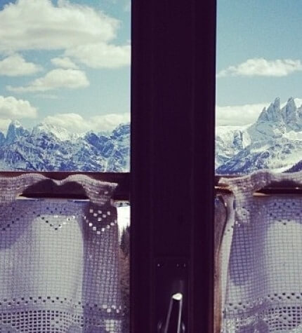 Scena serena di montagne innevate vista da una finestra con una tenda semi-trasparente e cielo blu con nuvole bianche.