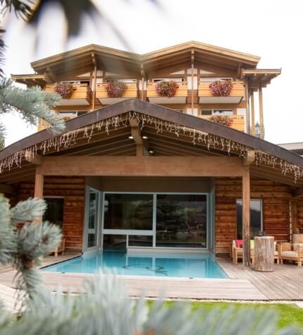 Edificio in legno a due piani con piscina interna, balcone fiorito e atmosfera serena, circondato da un cielo nuvoloso.