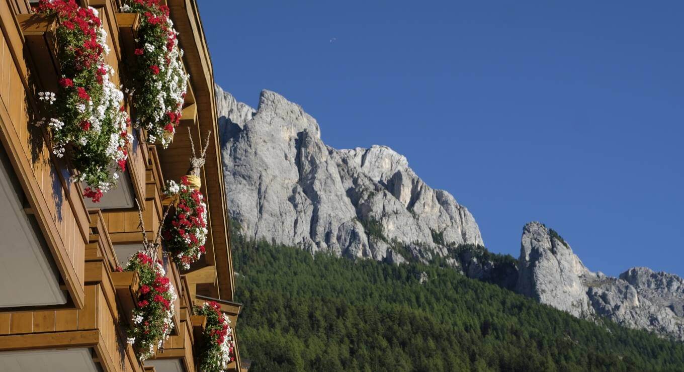 Paesaggio montano con un edificio in legno fiorito in primo piano e una maestosa catena montuosa sullo sfondo.