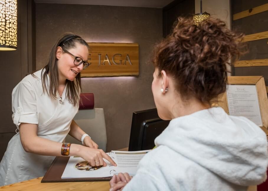 Donna in abito bianco parla con un'altra donna al tavolo, con un monitor e un'insegna "IAGA" sullo sfondo.