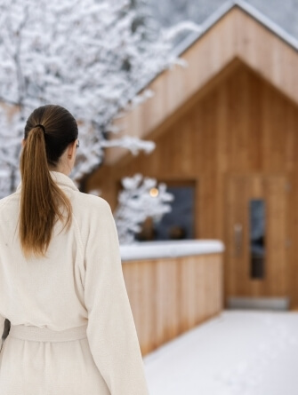 Donna in accappatoio bianco cammina verso un edificio di legno in un paesaggio innevato. Atmosfera invernale.