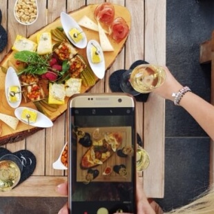 Le mani di una persona tengono uno smartphone mentre fotografano un tavolo di legno con cibo e bevande.