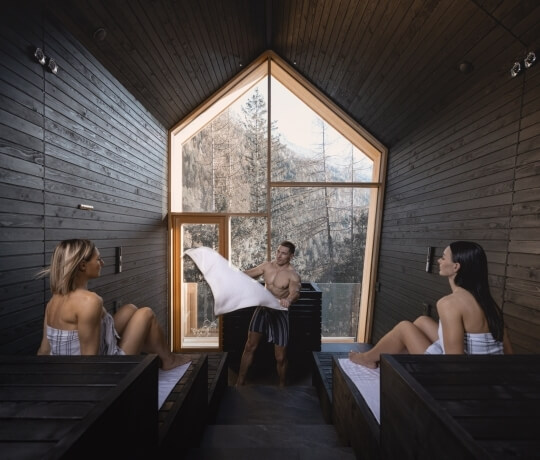 Donne su panche, uomo al centro, finestra su foresta e montagne.