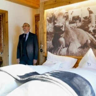 Un letto ordinato con una coperta a tema mucca, un uomo in giacca blu nella porta e una foto di mucca sul muro.