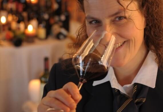 Donna con capelli ricci che annusa vino in un bicchiere, vestita con blazer nero e cravatta a righe gialle, in un ambiente formale.