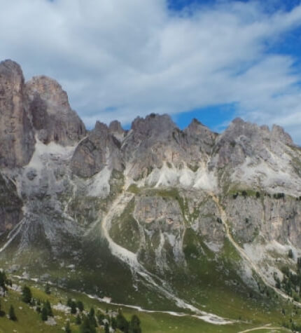 Catena montuosa maestosa con pendii ripidi, area verde lussureggiante in primo piano e cielo blu con nuvole bianche.