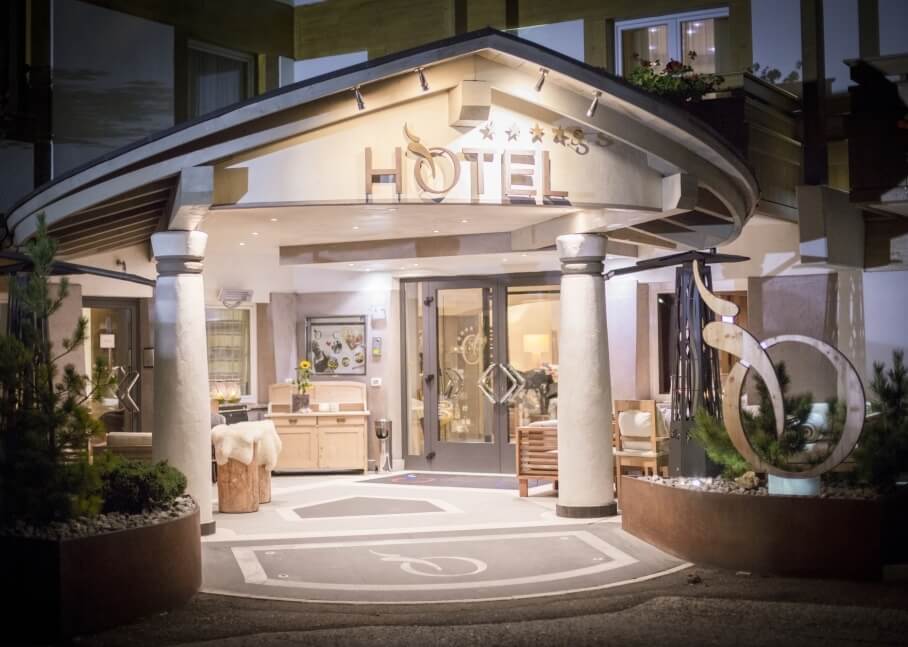 Ingresso di un hotel moderno con colonne bianche, insegna "HOTEL", piante e logo circolare, porte in vetro.