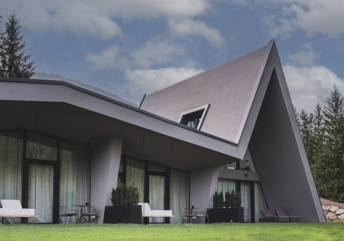 Casa moderna grigia con tetto angolare, portico coperto, grande prato e alberi, sotto un cielo blu con nuvole bianche.