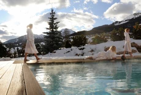 Tre persone in abiti bianchi camminano lungo un bordo piscina, con montagne innevate e cielo nuvoloso sullo sfondo.