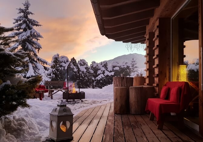 Una scena invernale serena con un'accogliente baita, un camino, una sedia rossa e un cielo colorato all'alba o al tramonto.