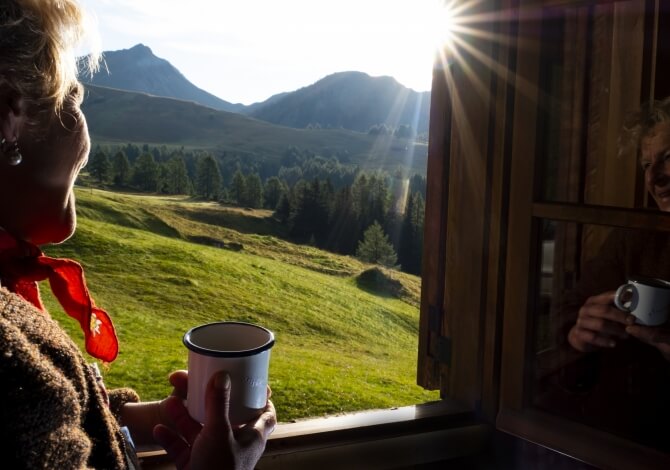 Una donna con capelli biondi guarda fuori dalla finestra, sorseggiando da una tazza, mentre un paesaggio sereno si estende oltre.