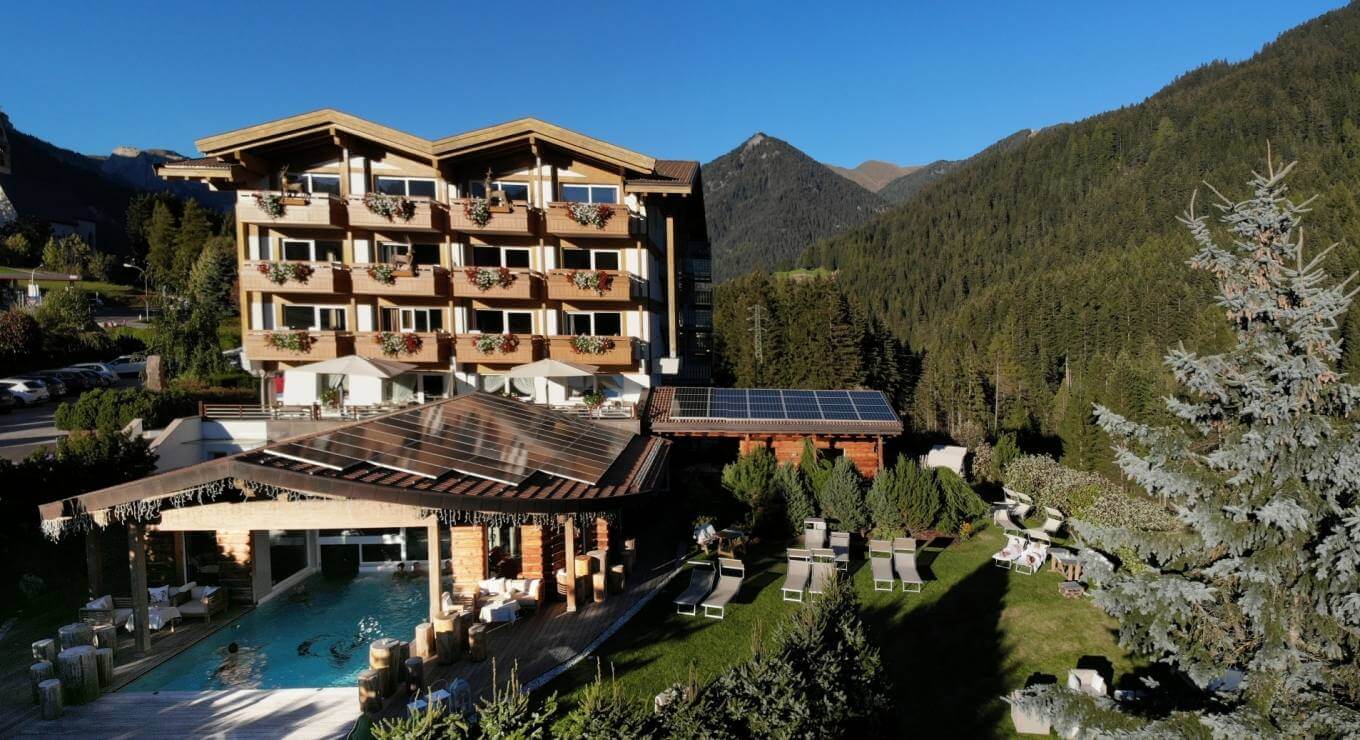 Edificio multi-storico in legno chiaro con balconi fioriti, piscina e montagne sullo sfondo, atmosfera di resort di lusso.