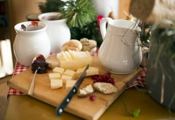 Tagliere di legno con formaggi, coltello e due brocche bianche, con un albero di Natale sullo sfondo. Atmosfera festiva.