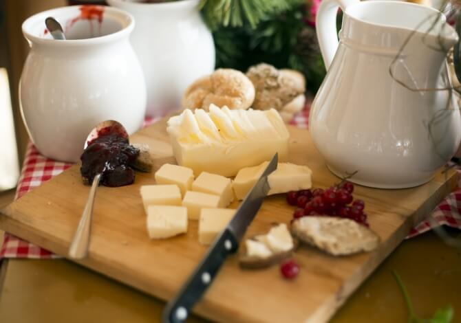 Tagliere di legno con formaggi, pane, ribes e marmellata, su tovaglia a quadretti rossi e bianchi, atmosfera festiva.