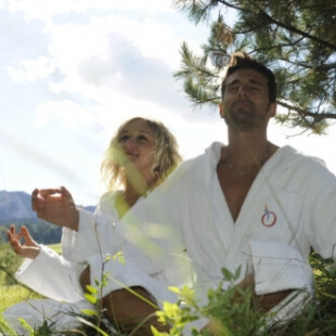 Uomo e donna in meditazione all'aperto, circondati da verde, montagne e cielo blu, con gesti di meditazione.