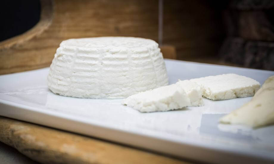 Un pezzo di formaggio bianco cilindrico su un piatto bianco, con due fette a destra e un altro formaggio sfocato in primo piano.