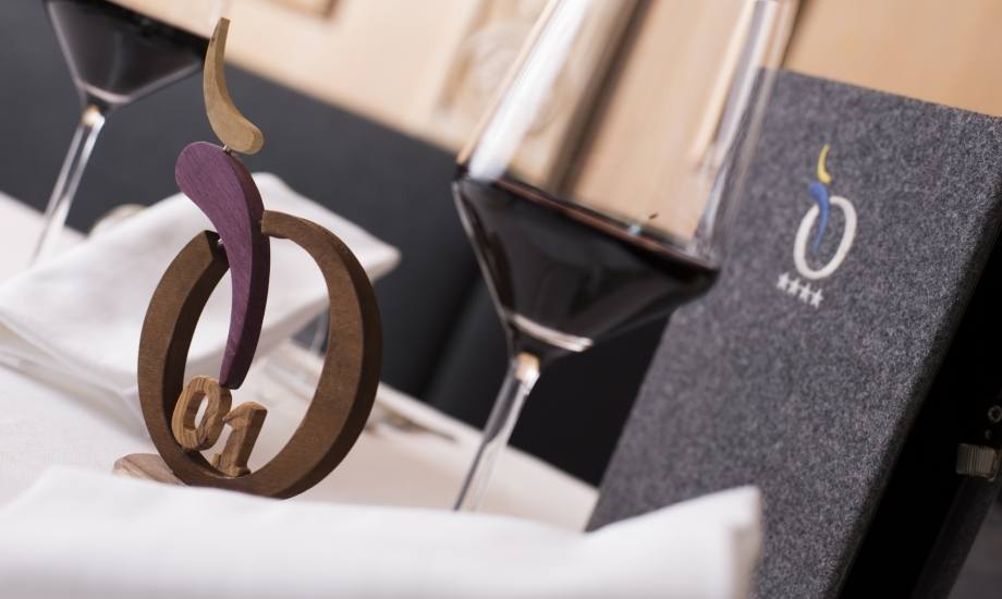 Impostazione della tavola con una scultura in legno colorato, un bicchiere di vino e un oggetto grigio con un logo.