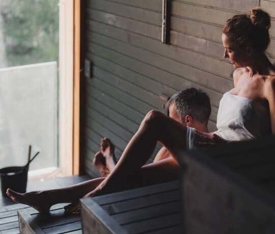 Due persone in una sauna di legno, avvolte in asciugamani bianchi.