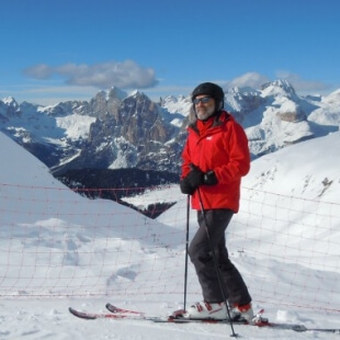 Uomo di mezza età che scia su una pista innevata, con montagne e cielo blu sullo sfondo.