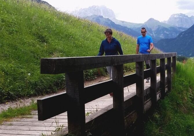 Due persone camminano su un ponte di legno, circondate da montagne e vegetazione lussureggiante in un'atmosfera serena.