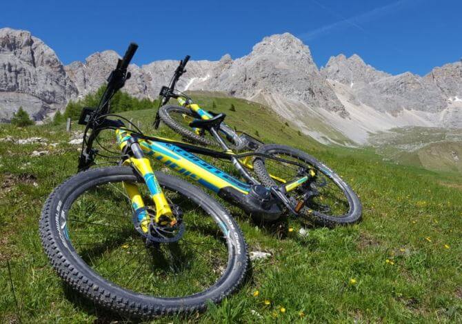 Due biciclette gialle con dettagli blu su una collina erbosa, con una catena montuosa sullo sfondo e cielo sereno.