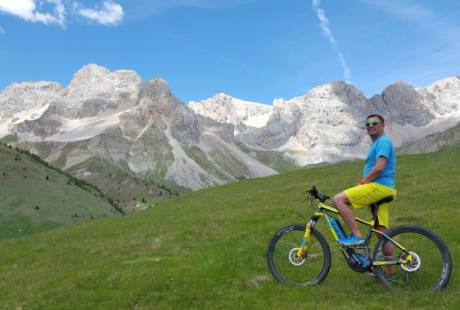 Un uomo in maglietta azzurra e pantaloni gialli pedala su una bici nera con dettagli gialli e blu in un prato verde con montagne.