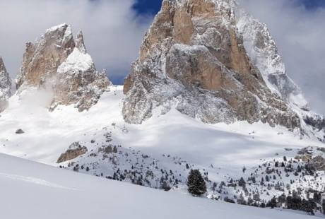 Paesaggio invernale sereno con montagne innevate, alberi sempreverdi e cielo blu con nuvole bianche.
