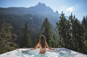 Una donna con capelli lunghi e bagnati si rilassa in una vasca idromassaggio, circondata da montagne e alberi verdi.