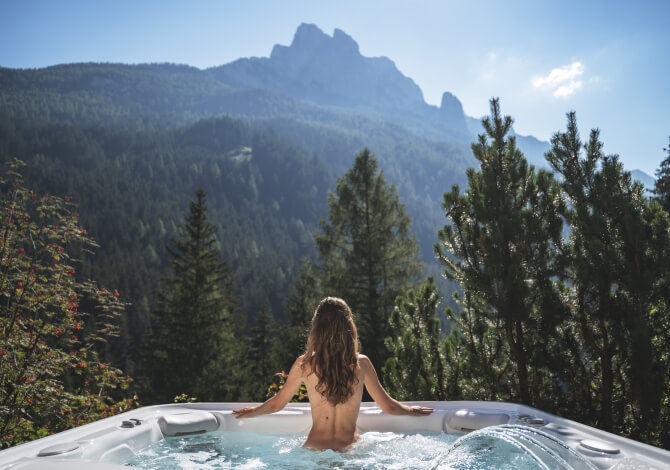 Una donna con lunghi capelli castani si rilassa in una vasca idromassaggio, circondata da montagne e vegetazione lussureggiante.