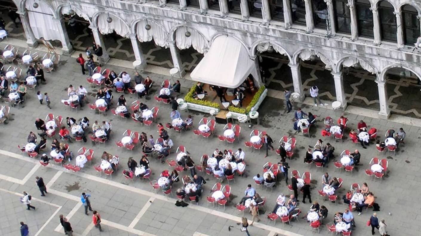 Vista aerea di un ristorante all'aperto con sedie rosse e tavoli bianchi, davanti a un edificio bianco con archi.