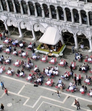 Area pranzo all'aperto con tavoli e sedie colorate davanti a un edificio storico, affollata di persone durante il giorno.