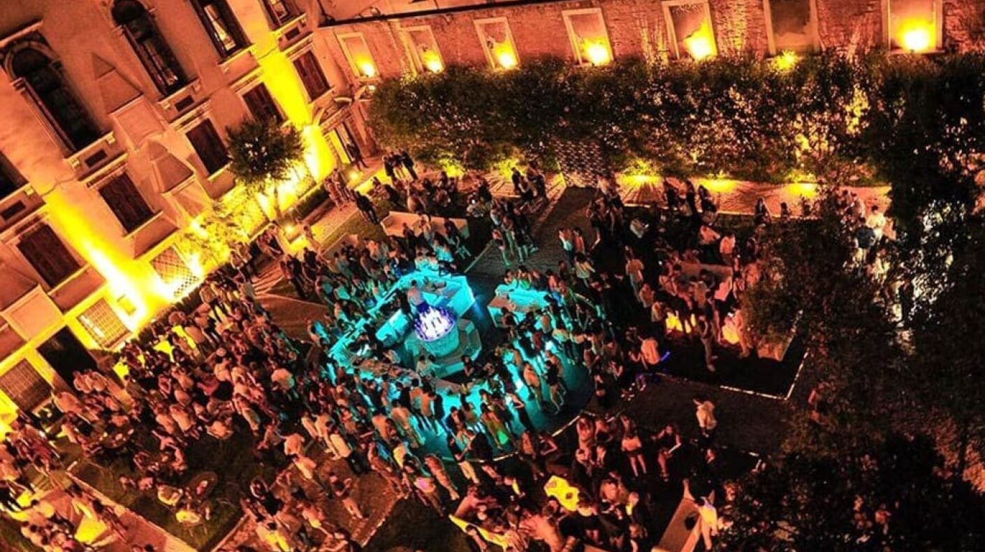 Raduno notturno vivace in un cortile, con una fontana blu e un edificio illuminato da luci gialle sullo sfondo.