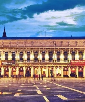Edificio architettonico simmetrico con finestre ad arco, cielo scuro e nuvoloso, strada bagnata in primo piano.