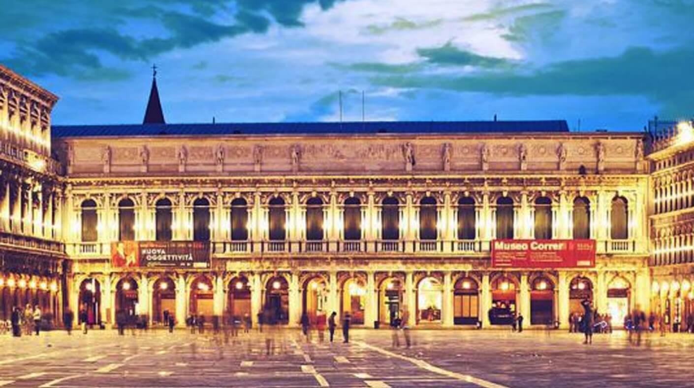 Edificio illuminato con facciata lunga, finestre ad arco e insegna "Museo Correr", con persone che passeggiano davanti.